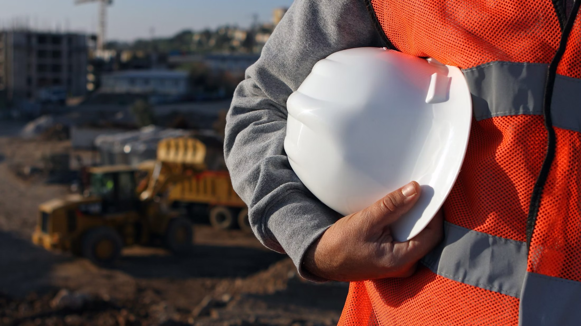 Bauarbeiter hält einen Schutzhelm, im Hintergrund eine Baustelle
